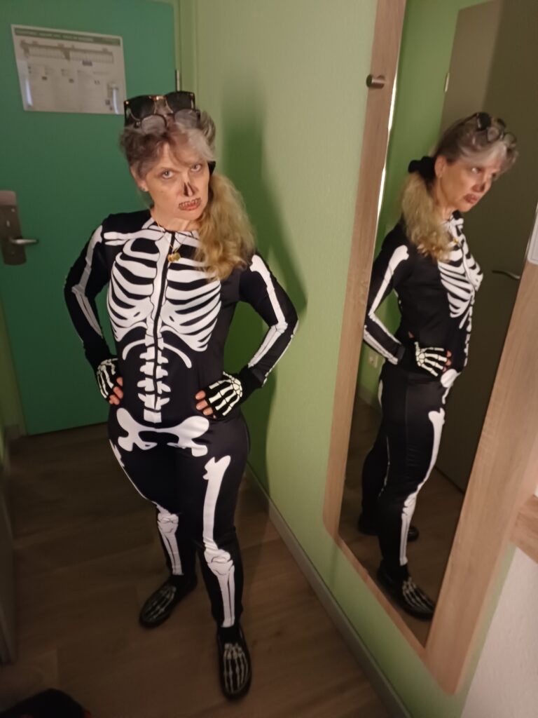 Frau mit Skelett-Bodysuit steht in einem Hotelzimmer neben einem Spiegel, damit man die Rückseite ebenfalls sehen kann. Das Gesicht ist andeutungsweise als Schädel geschminkt. Sie blickt düster in die Kamera, die Hände in die Hüfte gestemmt.