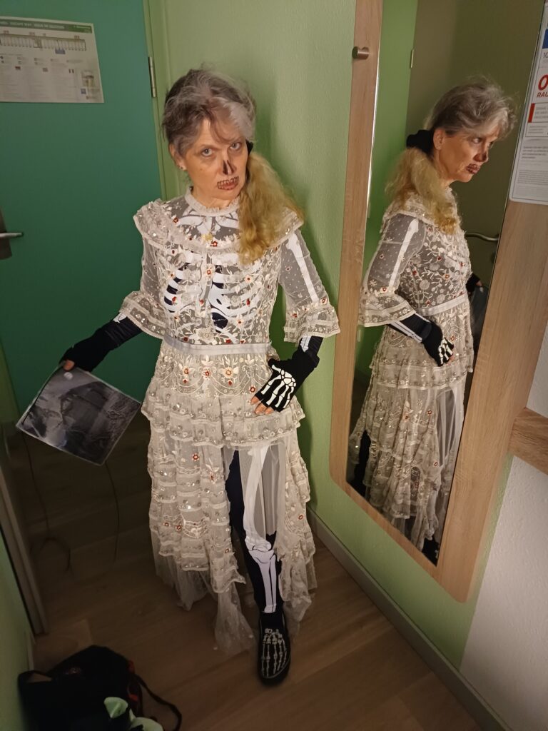 Dieselbe Frau in ähnlicher Pose, diesmal mit einem zerfledderten, altmodischen, weißen Kleid über dem Skelett-Bodysuit.