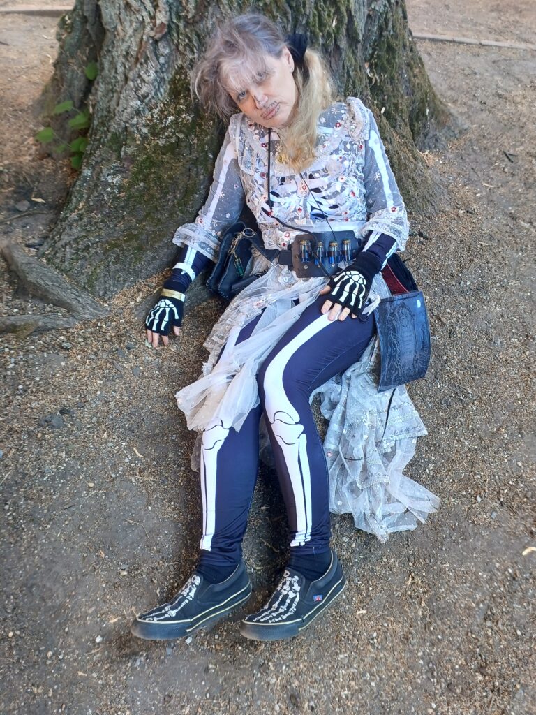 Frau im Skelett-Bodysuit und darüber ein zerfleddertes, altmodisches, weißes Kleid, das Gesicht andeutungsweise als Schädel geschminkt, liegt halb sitzend an einem Baumstumpf und blickt wehmütig in die Kamera.