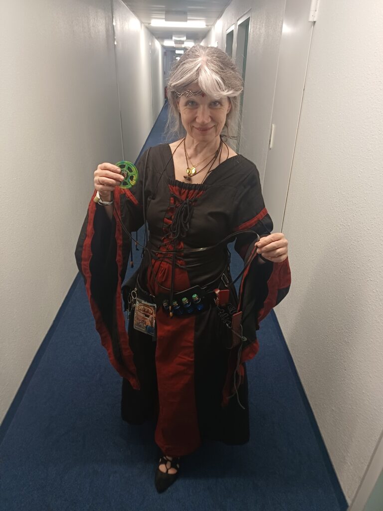 Frau mit schwarz-rotem Mittelalterkleid mit weiten Tubenärmeln und schwarzer Schnürung. Am Gürtel kleine Fläschchen mit blauer Flüssigkeit. Im silbernen Haar ein silberner Reif mit rotem Stein. In der Hand hält sie eine Rolle Super-8-Film. Sie steht in einem Hotelflur.