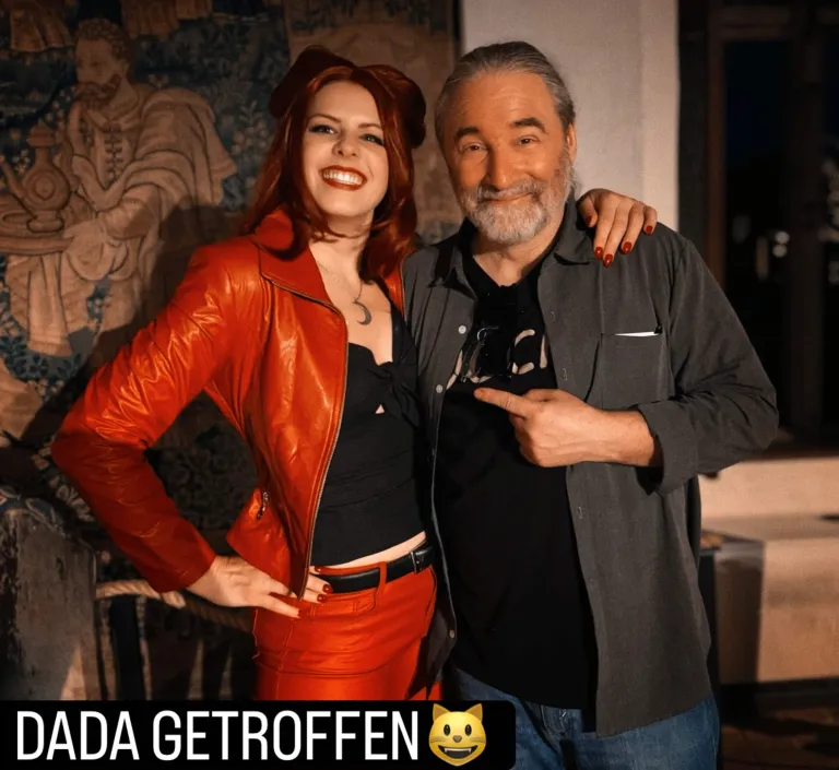 Tommy steht im DOCH.-Shirt und grauem Hemd neben einer Frau als Dada verkleidet, zeigt auf sie mit einem breiten Lächeln. Sie trägt rote Lederhosen mit schwarzem Gürtel, ein schwarzes Top, eine rote Lederjacke und eine rote Perrücke mit Katzenohren. Die Lippen sind ebenfalls rot, sie lächelt und strahlt, legt ihre Hand mit roten Fingernägeln auf Tommys Schulter, die andere in die Hüfte gestemmt. Darunter steht "Dada getroffen".