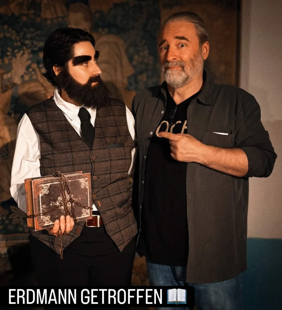 Tommy steht im DOCH.-Shirt und grauem Hemd neben einer Frau als Erdmann verkleidet, zeigt auf sie mit hochgezogenen Augenbrauen. Sie trägt sehr dichte künstliche Augenbrauen und einen ebensolchen Bart. Dazu schwarze Hose, weißes Hemd, schwarze Krawatte und eine braun-karierte Weste. In der Hand hält sie ein uraltes, braunes Buch, das mit einer Kette umwickelt ist. Darunter steht "Erdmann getroffen".