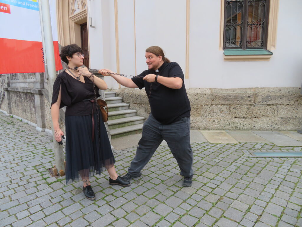 Frau mit schwarzem Kleid, Ledergürtel und Tasche, schwarzen Schuhen und schwarzen Fingerspitzen wird von einem Mann als Priester verkleidet in schwarzem Hemd an einer Kette um den Hals festgehalten. Sie legt Zeige- und Mittelfinger der linken Hand an ihren Hals und starrt den Mann an. Im Hintergrund ist die Treppe und der Eingang einer Kirche zu sehen.