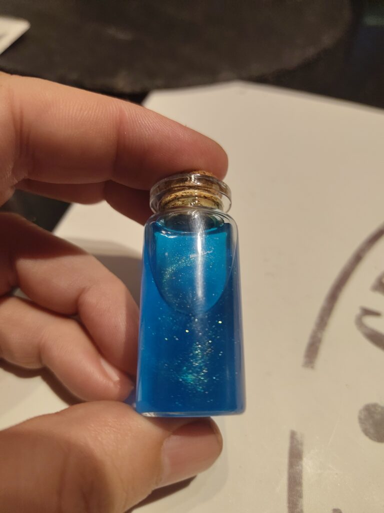 Kleines Glasfläschchen gefüllt mit blauer, glitzender Flüssigkeit mit einem Korken verschlossen. Das Fläschchen wird in einer Frauenhand gehalten.