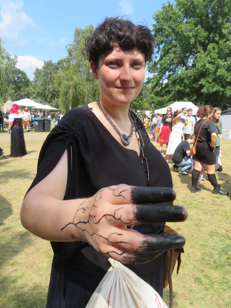 Eine Frau im schwarzen Kleid blickt geheimnisvoll in die Kamera. Ihre kurzen, braunen Haare sind gerkäuselt. Sie reckt eine Hand Richtung Kamera. Die Fingerspitzen sind schwarz gefärbt, auf dem Handrücken sind schwarze, verästelte Striche gemalt. Um den Hals trägt sie eine großgliedrige Kette und ein Halsband mit einem Metallring. Im Hintergrund sieht man das Treiben im Park während des Elbenwaldfestivals.