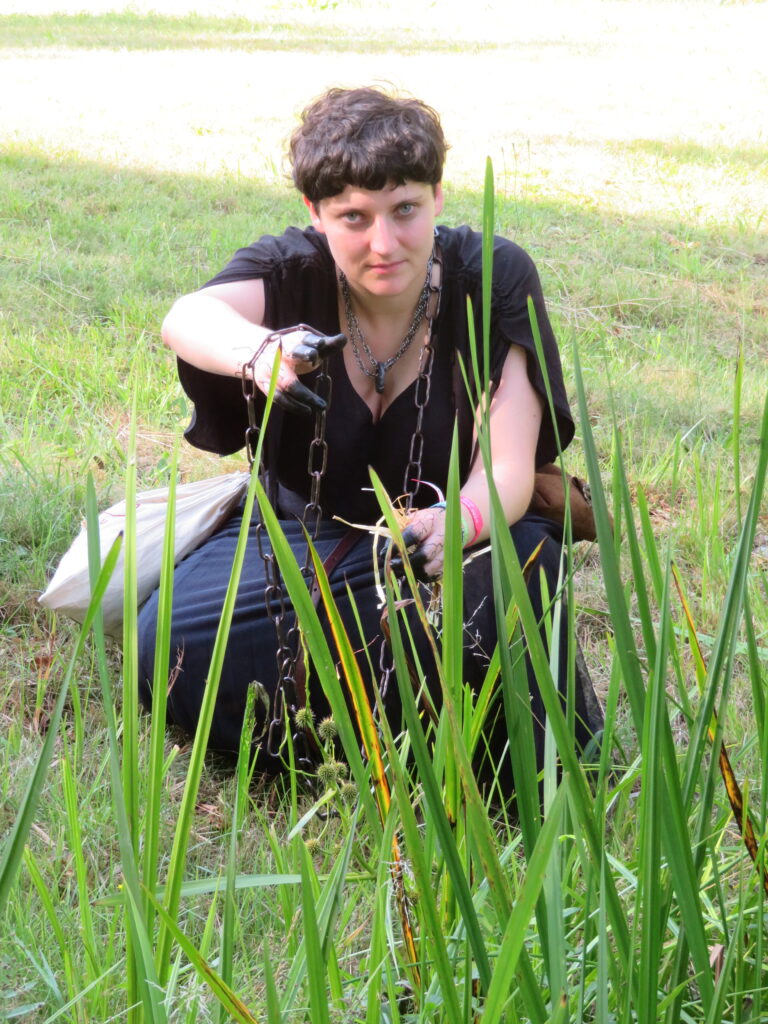 Im Gras hockt eine Frau mit scharzem Kleid. Ihre Fingerspitzen sind schwarz gefärbt und sie blickt finster in die Kamera. Um den Hals trägt sie eine Kette. Im Vordergrund stehen hohe Gräser. 
