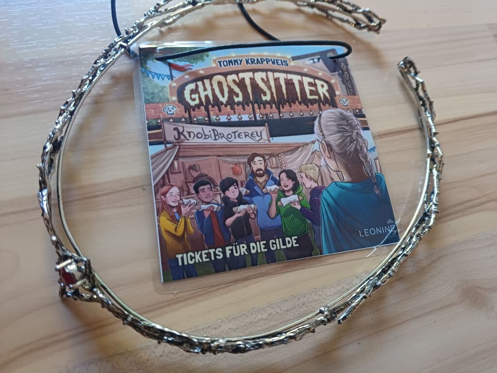 Silbernes Tiara mit rotem Stein liegt auf einem Parkettboden, in der Mitte das Titelbild der Ghostsitterfolge "Tickets für die Gilde", auf dem Mimi mit französischem Zopf zu sehen ist.